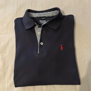 Marinblå pikétröja från Ralph Lauren - Passar Storlek S-M (har själv storlek S) Snygg marinblå pikétröja från Ralph Lauren med klassisk krage och knappar. Insidan av kragen och knappslån har rutigt mönster i svartvitt. Ikonisk röd logga broderad på bröstet. Perfekt för en stilren och avslappnad look.