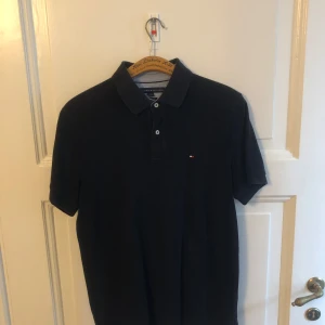 Svart piké från Tommy Hilfiger M - Klassisk svart pikétröja från Tommy Hilfiger i bomull. Custom fit-modell med korta ärmar, krage och knappar framtill. Liten broderad logga på bröstet. Perfekt för en clean och stilren look.