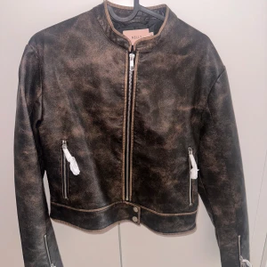 Vintage skinnjacka från Nelly XS - Snygg vintageinspirerad skinnjacka från Nelly i svartbrunt PU-material. Jackan har raka linjer, rundad krage, dragkedja framtill och dragkedjor vid fickor och ärmslut. Perfekt för dig som vill ha en edgy look med coola detaljer.