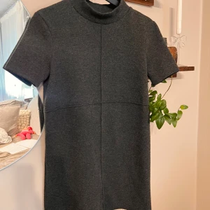 Mörkgrå kort klänning från Zara - Säljer en stilren mörkgrå klänning från Zara med korta ärmar och hög krage. Klänningen har en rak passform och är tillverkad i ett mjukt, stickat material som känns både bekvämt och trendigt. Perfekt för dig som gillar minimalistisk stil.