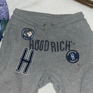 Grå Hoodrich shorts med tryck, strl L - Snygga grå mjukis från Hoodrich med stora tryck och broderade patches framtill. De har snörning i midjan och coola detaljer som logotyp på benet och etikett vid fickan. Perfekta för chill eller streetwear-stil. sitter som en M