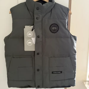 Canada Goose väst  - Exklusiv väst från Canada Goose Black Label i elegant grå färg. En lyxig och funktionell premiumväst som kombinerar värme och stil – perfekt för både vardag och fritid. 