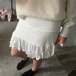 Vit kortkjol med volang - Supersöt vit kortkjol med bred smock i midjan och volangkant nedtill. Kjolen är gjord i ett mjukt och lätt tyg som ger en skön känsla och snygg passform. Perfekt för dig som gillar en feminin och fräsch stil.