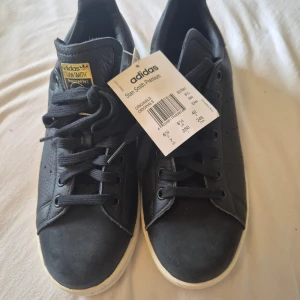 Adidas Stan Smith Premium svarta sneakers - Adidas Stan Smith Premium sneakers i svart skinn med klassisk rund tå och vit platt sula. Skorna har snörning, diskret perforering på sidorna och guldfärgade detaljer på plösen och innersulan. Perfekt för dig som gillar stilrena och tidlösa sneakers.