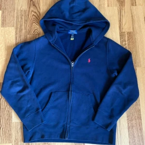 Marinblå hoodie från Polo Ralph Lauren - Säljer en marinblå zip-hoodie från polo Ralph lauren. Passar 165 till 175cm. Tröjan är i topp skick då den endast används en fåtal gånger. Tröjan har inga tecken på användning. Säljer på grund av att jag har växt ur den. Köpt för ca 1000kr. 