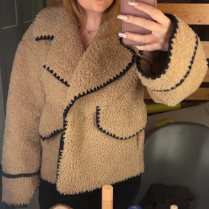 Beige teddyjacka med svarta detaljer - Supermysig beige teddyjacka med markerade svarta sömmar och stora slag. Jackan har två fickor framtill och oversized passform. Perfekt för att hålla sig varm och snygg under kyliga dagar. Köptes för 1299kr från object 