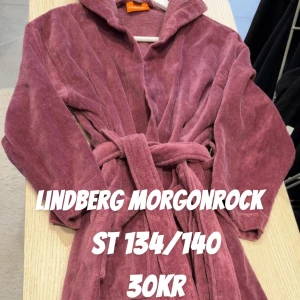 Lindberg lila morgonrock med huva - Mysig lila morgonrock från Lindberg med huva och knytskärp i midjan. Perfekt att slänga på sig efter duschen eller när du vill chilla hemma. Tillverkad i mjuk frotté och har långa ärmar för extra komfort.