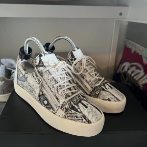 Giuseppe Zanotti sneakers i ormskinn - Säljer ett par sjukt snygga sneakers från Giuseppe Zanotti med ormskinnsmönster i svart och vitt. Skorna har snörning, dragkedjor på sidorna och en platt vit sula.                         Ovanlig modell samt i bra skick!