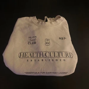 Vit sweatshirt från H&M Divided - Vit sweatshirt från H&M Divided med svart tryck framtill där det står 'HEALTHCULTURE ESTABLISHED' och flera andra grafiska detaljer. Tröjan har rund hals, ribbade muddar och är långärmad. Perfekt för en chill och sportig look.