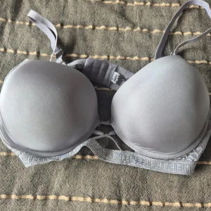 Silvergrå glittrig BH 85D - Snygg silvergrå BH med glittriga detaljer och spets på sidorna. Kuporna är släta och vadderade för en snygg form. Axelbanden är justerbara och bandet har glittrande stenar som ger extra bling. Perfekt för dig som vill ha något extra under kläderna.