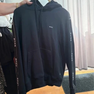 Svart hoodie från BOSS med logga - Svart hoodie från BOSS med huva och snörning. Tröjan har logga på bröstet och längs ärmarna, samt en stor magficka. Perfekt för en chill och stilren look.