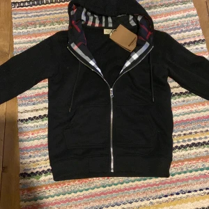 Svart Burberry hoodie med dragkedja - Svart hoodie från Burberry med klassiskt rutigt foder i huvan och längs insidan, dragkedja framtill och snörning vid huvan. Tröjan har två fickor och långa ärmar. Perfekt för dig som gillar stilrena designer med en touch av lyx.