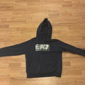Svart EA7 hoodie med logga - Svart hoodie från EA7 Emporio Armani med stor silverfärgad logga på bröstet. Tröjan har huva, lång ärm och en klassisk magficka framtill. Perfekt för dig som gillar stilrena streetwear-plagg med exklusiv känsla.
