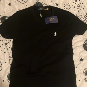 Svart t-shirt från Polo Ralph Lauren - Svart t-shirt från Polo Ralph Lauren i storlek S. Klassisk design med vit broderad logga på bröstet och rund hals. Tillverkad i mjuk bomull som känns skön mot huden. Perfekt för dig som gillar stilrena och tidlösa plagg med premiumkänsla.