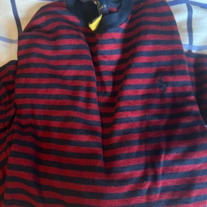Randig tröja från Polo Ralph Lauren - Snygg randig tröja från Polo Ralph Lauren i mörkblått och rött. Tröjan har klassisk rund hals och diskret broderad logga på bröstet. Perfekt för dig som gillar stilrena och tidlösa plagg med en sportig touch.