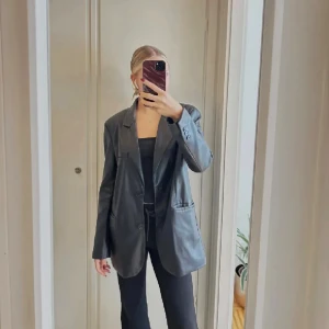 Svart oversized skinnkavaj - Säljer en svart oversized kavaj i skinnimitation med klassisk krage och knäppning framtill. Jackan har långa ärmar, två fickor och en clean look som ger en cool vibe. Perfekt för dig som vill ha en edgy och trendig stil.