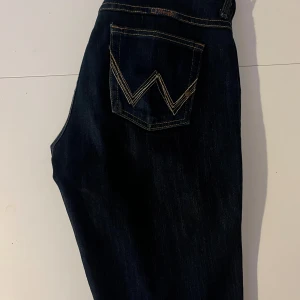 Wrangler  mörkblå jeans med broderad ficka - Snygga mörkblå jeans från wrangler med klassisk femficksdesign och coolt broderad W på bakfickan. Jeansen har guldfärgade sömmar, dragkedja och knapp framtill samt ett mönstrat midjeband på insidan. Perfekt passform och stilren look.