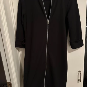 Svart Boohoo klänning med dragkedja strl: 36 - Svart kortklänning från Boohoo med snygg silverfärgad dragkedja framtill och trekvartsärmar med uppvik. Stilren och enkel design i mjukt syntetmaterial, perfekt för dig som gillar minimalistiskt och cleant uttryck.