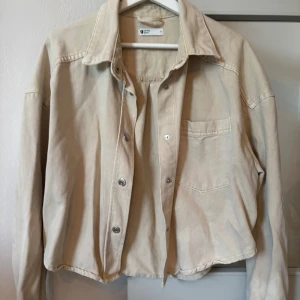 Beige jeans jacka från Gina Tricot - Snygg beige overshirt från Gina Tricot med klassisk krage, bröstficka och silverfärgade knappar. Jackan är i bomull och har en avslappnad passform som är perfekt att slänga över en t-shirt. Långärmad och enkel att styla till det mesta.