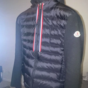 Svart Moncler dunjacka med huva - Svart dunjacka från Moncler med quiltad front och huva. Jackan har stickade ärmar, dragkedja framtill och röda/vita detaljer på snörningen. Klassisk Moncler-logga på ärmen och snygg, sportig look. Perfekt för kyliga dagar. Det står storklek M men det passar S och även XS. Pris kan diskuteras.