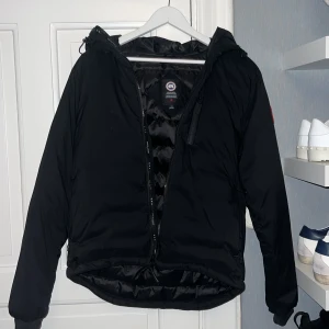 Svart dunjacka från Canada Goose M - Säljer en svart dunjacka från Canada Goose med huva och dragkedja. Jackan har klassisk logga på ärmen, flera fickor och en stilren, sportig look. Perfekt för kalla dagar och riktigt snygg till streetwear. Passar dig som vill ha både värme och stil. Ny pris 9400kr