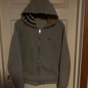 Grå hoodiejacka från Burberry - Säljer en grå hoodiejacka från Burberry med klassiskt rutigt foder i beige, svart och rött. Jackan har dragkedja, två fickor fram och broderad Burberry-logga på bröstet. Perfekt för dig som gillar stilren design med lyxig touch. Tröjan har en skada ner vid kedjan men går lätt att sy ihop, tröjan har inga snören till luvan.