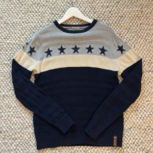 Stickad tröja med stjärnor från Hilfiger - Stickad tröja från Hilfiger Denim i blått, grått och beige. Tröjan har ett mönster med mörkblå stjärnor över bröstet och ribbade muddar vid ärmslut och nederkant. Perfekt för dig som gillar en klassisk men ändå unik look.