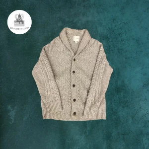 Grå stickad kofta från American Eagle - Säljer en grå stickad kofta från American Eagle med snyggt kabelstickat mönster och stora knappar framtill. Koftan har bred krage och långa ärmar, perfekt för lager-på-lager. Skön och stilren att slänga på sig när det är kyligt ute. Jackan är L storlek men passar till en större M. Har en liten fläck på insidan som inte synns utifrån