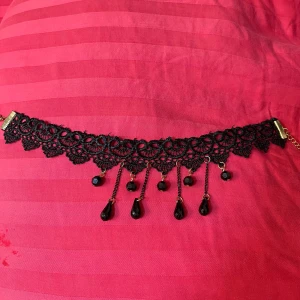 Svart spetschoker med pärlor - Unik svart choker i spets med dekorativa mönster och hängande svarta pärlor och droppformade hängen. Kedjor och detaljer i guldfärg ger extra edge. Perfekt för dig som gillar statement-smycken med gotisk vibe.
