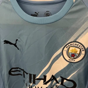 Manchester City matchtröja Puma blå - Manchester Citys officiella matchtröja från Puma i ljusblått med vita detaljer och klubbmärke på bröstet. Tröjan har korta ärmar, rund hals och är tillverkad i ett lätt, ventilerande funktionsmaterial. Snyggt tryck med Etihad Airways på framsidan.