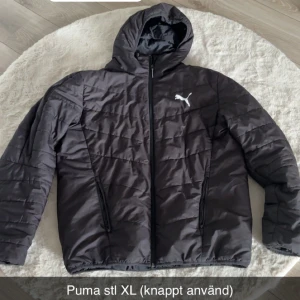 Svart pufferjacka från Puma XL - Svart pufferjacka från Puma i storlek XL med huva och dragkedja framtill. Jackan har två snedställda fickor med dragkedja och ett vitt Puma-logo på bröstet. Perfekt för kyligare dagar och enkel att matcha med det mesta.