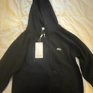 Svart hoodie från Lacoste med dragkedja - Svart hoodie från Lacoste med klassisk krokodillogga på bröstet. Tröjan har huva med snörning, hel dragkedja framtill och långa ärmar. Tillverkad i mjuk bomullsmix som är perfekt för höst och vinter. Snygg och enkel design som funkar till allt.