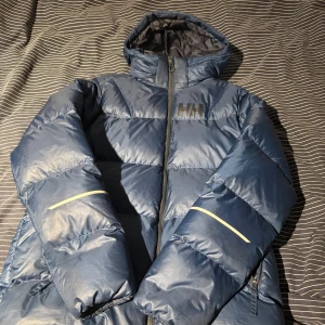 Blå pufferjacka Helly Hansen  - Blå pufferjacka från Helly Hansen med huva och dragkedja. Jackan har reflekterande detaljer på ärmarna och HH-logga på bröstet. Tillverkad i syntetmaterial och fodrad för extra värme. Perfekt för kalla dagar när du vill hålla stilen.