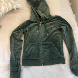 Grön velour hoodie Juicy Couture XS - Mjuk och lyxig grön hoodie från Juicy Couture i velour med dragkedja framtill, huva med snörning och känguruficka. Klassisk logga broderad på bröstet. Perfekt för en chill och trendig look.