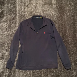 Mörkblå quarter zip Polo Ralph Lauren - Säljer en mörkblå långärmad tröja från Polo Ralph Lauren med röd broderad logga på bröstet. Tröjan har en snygg half zip-dragkedja och klassisk krage. Tillverkad i mjuk bomull, perfekt för lager-på-lager under höst och vinter. Storlek M