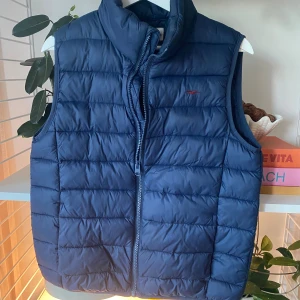 Blå dunväst från Hampton Republic - Snygg marinblå dunväst från Hampton Republic med quiltad design och dragkedja framtill. Västen har en liten röd broderad logga på bröstet och hög krage. Perfekt för lager-på-lager och enkel att matcha med hoodies eller tröjor.