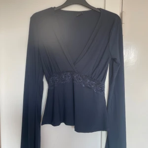 Mörkblå omlottopp med spets Gina Tricot - Supersnygg mörkblå omlottopp från Gina Tricot i stretchigt material. Toppen har långa ärmar, v-ringning och en bred spetsdetalj med blommönster i midjan. Perfekt för dig som gillar feminina detaljer och en figurnära passform. Säljer för att den är för stor 💐🌸