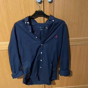 Mörkblå skjorta från Ralph Lauren - Mörkblå skjorta från Ralph Lauren i slim fit med klassisk krage och vita knappar. Broderad röd logga på bröstet. Tillverkad i lätt bomullstyg, perfekt för en clean och stilren look. Kommer strykas innan den skickas. Hör av dig vid minsta fundering!