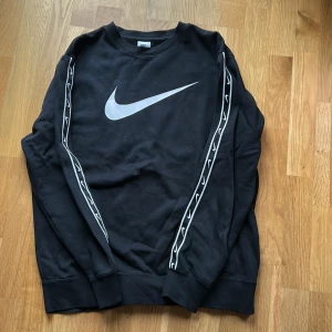 Svart Nike sweatshirt med logga - Svart sweatshirt från Nike med stor vit swoosh-logga på bröstet och vita loggor längs båda ärmarna. Klassisk rund halsringning och ribbade muddar. Perfekt för en sportig och avslappnad stil.