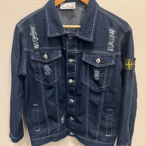 Stone Island blå jeansjacka med slitningar - Snygg blå jeansjacka från Stone Island med slitna detaljer och tydliga vita sömmar. Klassisk modell med bröstfickor, knappar framtill och ikonisk patch på ärmen. Perfekt för dig som vill ha en streetig och trendig look.