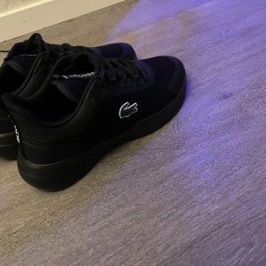 Svarta sneakers från Lacoste - Säljer ett par svarta sneakers från Lacoste med klassisk krokodillogga på sidan. Skorna har snörning, rund tå och platt sula. Ovandelen är i syntetmaterial och textil, vilket ger en clean och sportig look. Perfekta för dig som gillar stilrena och bekväma skor.