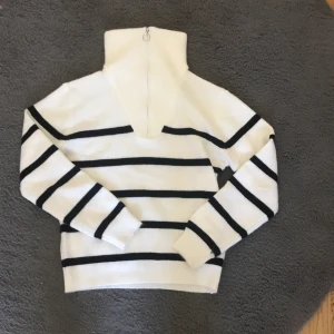Vit och svart randig stickad tröja - Snygg vit stickad tröja med svarta horisontella ränder och hög krage med silverfärgad half zip-dragkedja. Tröjan har långa ärmar och ribbade muddar. Perfekt för lager-på-lager och ger en fräsch, klassisk look.