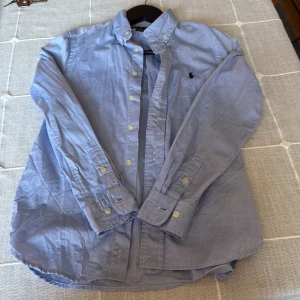 Blå skjorta från Ralph Lauren - Klassisk ljusblå skjorta från Ralph Lauren med button down-krage och broderad logga på bröstet. Skjortan har långa ärmar, knappar framtill och är tillverkad i mjuk bomull. Perfekt för en clean och stilren look. Det är storlek 12 år på denna fina skjorta och är i slim fit har använt denna 1 gång och inget mer använde denna till en speciell fest och nu efter ett tag har den blivit liten för mig den är i nytt skick ny pris ca 1100 mitt pris bara 500kr