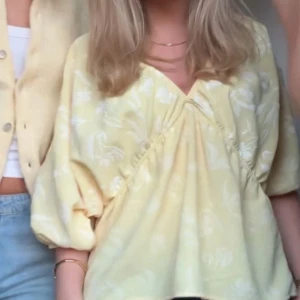 Gul blommig blus med omlott och puffärm - Säljer en ljusgul blus med vitt blommigt mönster och omlottdetalj framtill. Blusen har puffiga trekvartsärmar och en v-ringad hals. Perfekt för dig som gillar romantisk och fräsch stil med lite extra volym i ärmarna.