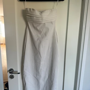 Vit långklänning - Snygg vit långklänning och stilren, veckad överdel. Klänningen har en figurnära passform och är axelbandslös upptill, vilket ger en elegant siluett. Perfekt för dig som gillar minimalistisk och klassisk stil. Den går ner till vaderna på mig som är 170. Den är utan band. Hänger på galghängarna.