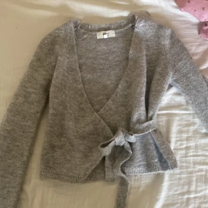 Grå omlottopp från Gina Tricot - Mysig grå omlottopp från Gina Tricot i ullblandning. Toppen har långa ärmar, knytband i midjan och en croppad passform. Perfekt för lager på lager och enkel att styla med olika outfits.