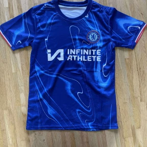 Chelsea FC blå matchtröja 2023/24 - Chelsea FCs officiella blå matchtröja för säsongen 2023/24 med vit logga och Infinite Athlete-tryck på bröstet. Tröjan har korta ärmar med röda och vita detaljer vid ärmslut, samt ett unikt abstrakt mönster i olika blå nyanser. Tillverkad i lätt och ventilerande polyester.