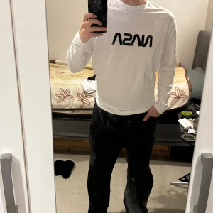 Vit långärmad NASA-tröja - Vit långärmad tröja med svart NASA-tryck på bröstet och stor färgglad jordglobsgrafik samt flera NASA-loggor på ryggen. Skön passform och tillverkad i mjuk bomull, perfekt för dig som gillar rymd och coola prints.