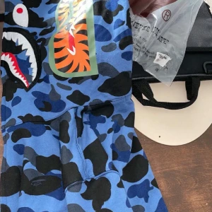 BAPE blå camo shark hoodie - Säljer en ikonisk blå camo shark hoodie från BAPE med hel dragkedja som går upp över huvan. Tröjan har det klassiska hajansiktet broderat på huvan och färgstarka detaljer. Perfekt statement-plagg för streetwear-entusiasten.
