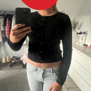 Svart kofta med knappar - Säljer en svart, croppad kofta med rund hals och knäppning framtill. Tröjan har lång ärm och sitter snyggt figurnära. Perfekt att styla med jeans eller kjol för en clean look.